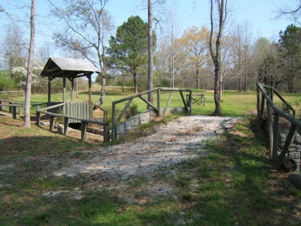 A serene outdoor scene featuring a wooden bridge leading to a grassy area, surrounded by trees. To the left, there is a small shelter with a peaked roof and a bench, while a rustic wooden fence runs alongside the path. The sky is clear and blue, creating a tranquil atmosphere. Georgia International Horse Park mountain bike trail.