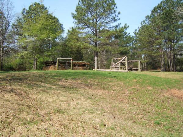 A grassy hillside with scattered trees in the background. On the left, a wooden structure resembling a shelter is partially visible, while on the right, an old wooden gate stands open, leading to a pathway. The scene is set on a sunny day with clear blue skies. Georgia International Horse Park mountain bike trail.