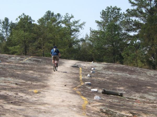 A person riding a mountain bike along a rugged trail on a rocky surface, bordered by tall trees in the background. The path is marked with yellow lines, and there are small rocks and logs scattered along the trail. Georgia International Horse Park mountain bike trail.
