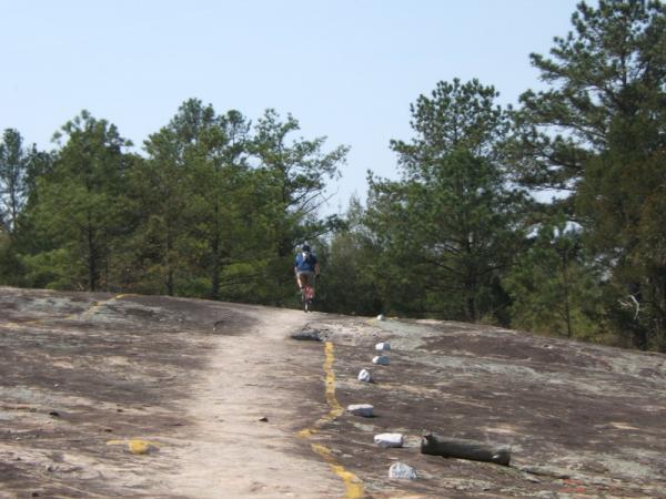 A person riding a bicycle along a rocky path surrounded by trees under a clear sky. The trail is marked with yellow lines, leading up a slope. Georgia International Horse Park mountain bike trail.