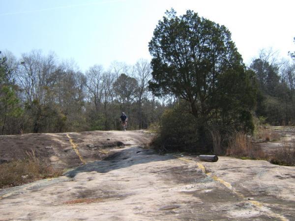 A person walking on a rocky trail surrounded by sparse trees on a sunny day. The landscape features exposed rock formations and scattered vegetation. Georgia International Horse Park mountain bike trail.