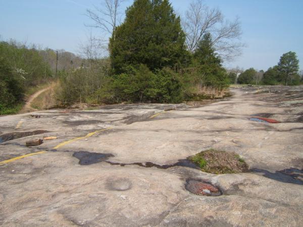 A rocky, flat landscape with patches of greenery and trees in the background, featuring a smooth stone surface. Yellow markings are visible on the rock, indicating a former road. In the distance, a dirt path winds through the trees. The scene is set under a clear blue sky. Georgia International Horse Park mountain bike trail.