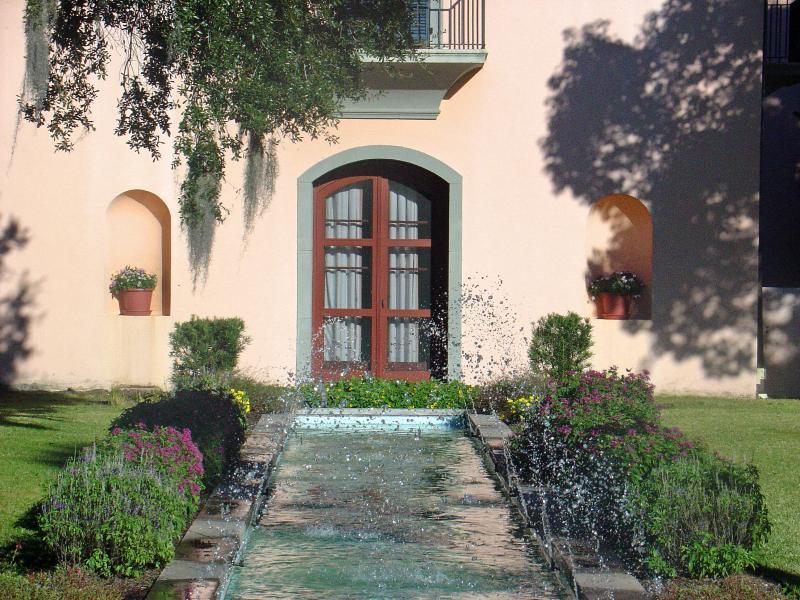 A tranquil scene featuring a fountain in a landscaped garden, with a pathway of water surrounded by colorful flowers. In the background, a pale pink building with large, wooden double doors and two arched windows is visible. Potted plants are situated near the lower windows, completing the serene garden setting. Family Fun mountain bike trail.
