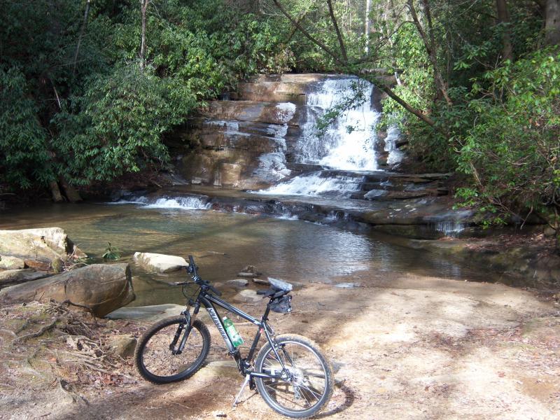 A mountain bike resting on the shore of a serene stream, with a waterfall cascading over rocks in the background and surrounded by lush greenery. Stonewall Falls Loop mountain bike trail.