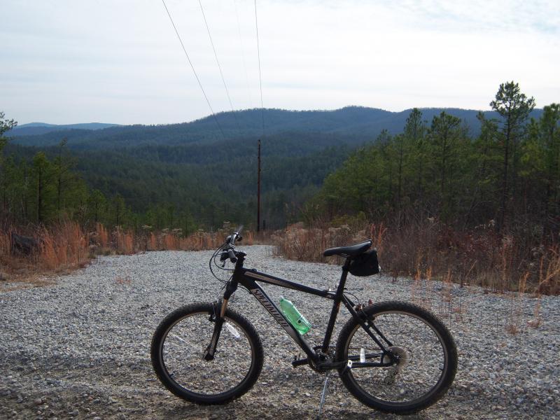 A mountain bike with a water bottle is parked on a rocky path, overlooking a scenic vista of rolling hills and forested terrain under a cloudy sky. Power lines are visible in the background. Stonewall Falls Loop mountain bike trail.