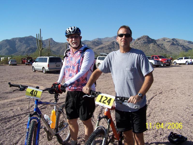 Two men stand next to their mountain bikes at a desert location. One man is wearing a helmet and colorful cycling attire, while the other is dressed in a gray t-shirt and shorts. Both bikes have numbered tags prominently displayed: one showing "40" and the other "124." In the background, there are vehicles parked, and the landscape features mountains and cacti under a clear blue sky. The date displayed in the corner reads November 4, 2006. Hawes Loop mountain bike trail.