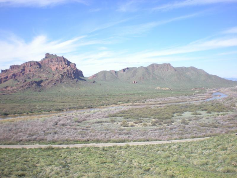 A panoramic view of a desert landscape featuring rugged mountains, a meandering river, and sparse vegetation under a blue sky with wispy clouds. The foreground includes a winding path and patches of dry grass and shrubs, while the background showcases rocky formations and rolling hills. Hawes Loop mountain bike trail.