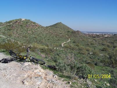 A mountain bike resting on rocky terrain with a sprawling green landscape and hills in the background. A dirt path winds through the scenery, under a clear blue sky. The image is dated January 5, 2004. Hawes Loop mountain bike trail.