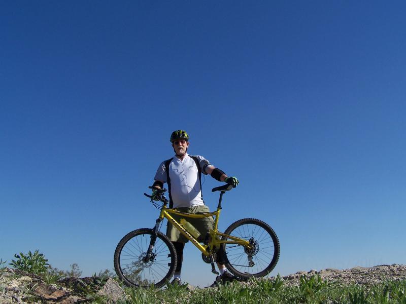 A cyclist stands proudly with a yellow mountain bike, posing against a clear blue sky. He wears a helmet and cycling gear, exuding enthusiasm for outdoor adventure. Lush greenery can be seen in the foreground, framing the scene. Hawes Loop mountain bike trail.
