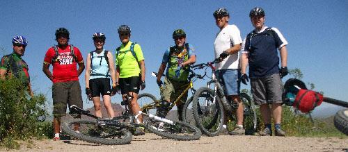 A group of seven mountain bikers poses for a photo on a sunny day, with their bikes resting on the ground beside them. They are wearing helmets and a mix of colorful cycling attire, standing against a clear blue sky. Hawes Loop mountain bike trail.