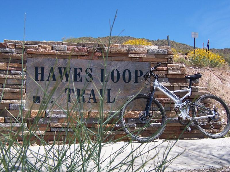A mountain bike leaning against a stone sign that reads "Hawes Loop Trail," surrounded by desert vegetation and a clear blue sky. Hawes Loop mountain bike trail.