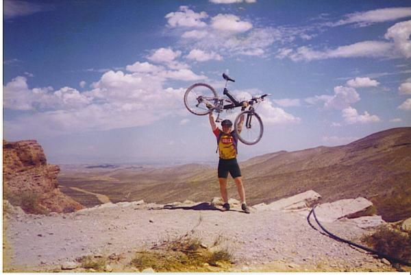 A person standing on a rocky outcrop in a desert landscape, joyfully holding a mountain bike above their head. The sky is filled with fluffy clouds and the scene captures a sense of accomplishment and adventure. Cottonwood Valley North mountain bike trail.