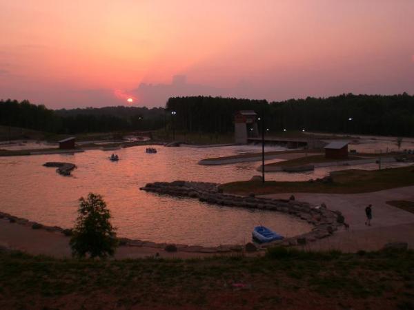 Sunset view over a tranquil lake with rocky edges, surrounded by greenery. Small boats are on the water, and several wooden structures are visible along the shoreline. The sky is painted in shades of orange and pink as the sun sets in the background. USNWC mountain bike trail.