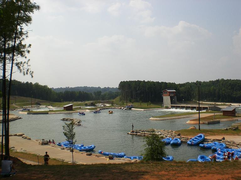 A scenic outdoor view of a water activity area featuring a calm lake surrounded by lush green trees. Blue inflatable rafts are lined along the shore, with some people enjoying the water and others walking on the dock. A small building with a tower overlooks the area, and the sky is partly cloudy. USNWC mountain bike trail.