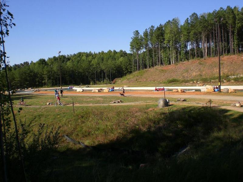 A dirt racing track surrounded by trees, with people riding go-karts and walking along the path. The scene includes natural greenery and rocks scattered around the track, under a clear blue sky. USNWC mountain bike trail.