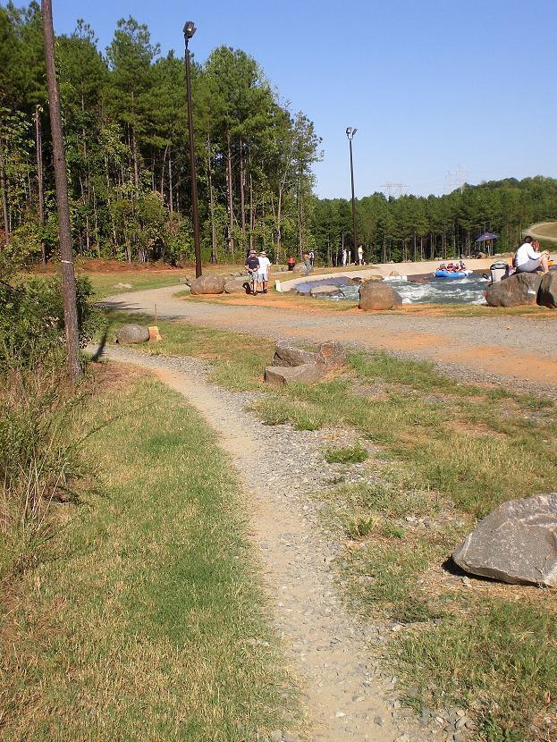 A scenic outdoor path lined with grass and rocks, leading towards a water feature. In the background, groups of people are enjoying leisure activities near the water, surrounded by tall trees under a clear blue sky. USNWC mountain bike trail.