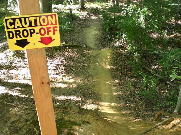 A yellow caution sign reading "CAUTION DROP-OFF" is mounted on a wooden post near a narrow dirt path winding through a wooded area. The path slopes downwards, surrounded by trees and scattered leaves, indicating a drop-off ahead. USNWC mountain bike trail.