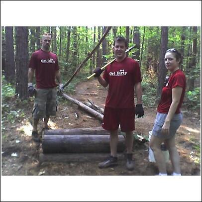 Three individuals are standing in a wooded area, surrounded by tall green trees. They are wearing matching red t-shirts with the phrase "Get Dirty" printed on them. The man on the left is dressed in camouflage shorts, while the man in the center, who holds a tool, is wearing red shorts. The woman on the right is in casual attire with a ponytail. They appear to be involved in outdoor work, with logs laid out on the ground behind them. USNWC mountain bike trail.