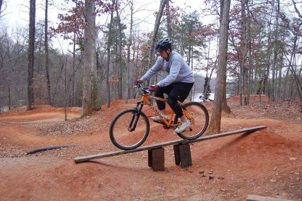 A person wearing a helmet and gray sweatshirt rides an orange mountain bike over a wooden plank bridge in a dirt trail park surrounded by trees. The terrain features dirt mounds and is partially cleared, indicating a biking or BMX track. Country Park mountain bike trail.