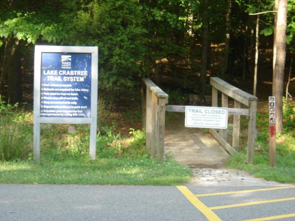 Sign for Lake Crabtree Trail System with a closed trail notice. The image shows a wooden entrance to a trail surrounded by trees, with two signs: one detailing park rules and the other indicating the trail is closed. Lake Crabtree County Park mountain bike trail.