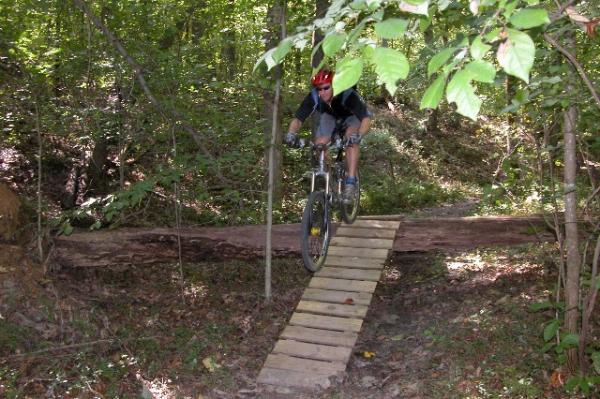 A mountain biker rides over a narrow wooden bridge set in a wooded area, surrounded by lush green trees and foliage. Legend Park mountain bike trail.