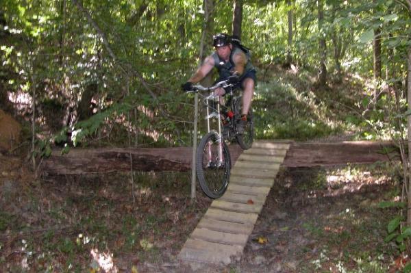 A mountain biker in a sleeveless shirt and shorts is mid-jump over a wooden bridge in a wooded area. The background features dense trees and underbrush, indicating a natural trail. The biker is wearing a helmet and appears focused as he navigates the challenging terrain. Legend Park mountain bike trail.