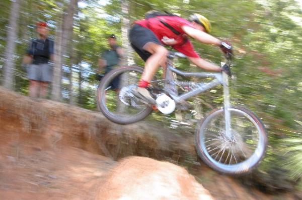 A mountain biker in a red shirt and helmet is mid-air, jumping off a rocky ledge on a forest trail. Two spectators in the background watch as the biker navigates the rugged terrain. The image captures a dynamic moment of action in a wooded environment. Legend Park mountain bike trail.