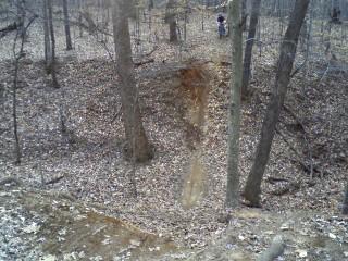 A wooded area with trees and fallen leaves covering the ground. In the foreground, there is a small erosion feature or slope leading down into a depression. A person can be seen in the background, near the top of the slope. Owls Roost (Bur-Mil Park) mountain bike trail.