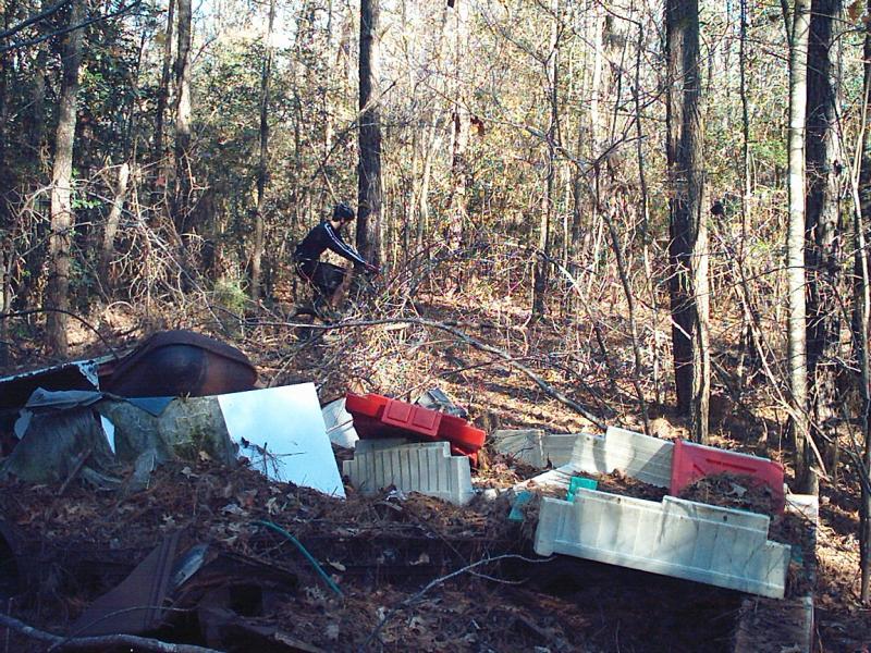 A person wearing a black outfit is navigating through a wooded area filled with debris and discarded items, including plastic containers and scrap metal. The scene is characterized by trees in the background and a cluttered foreground, suggesting an area that has been affected by littering or abandonment. Bicycle Post Trails mountain bike trail.