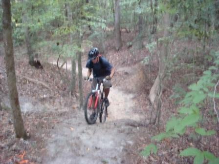 A person riding a mountain bike uphill through a wooded trail, surrounded by trees and natural foliage. Bicycle Post Trails mountain bike trail.