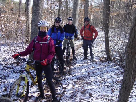 Four mountain bikers pose on a snowy trail in a forest. They are dressed in winter cycling gear, including helmets and gloves. The terrain is partially covered in snow, and trees surround the scene, indicating a cold, crisp day for riding. Reedy Fork mountain bike trail.