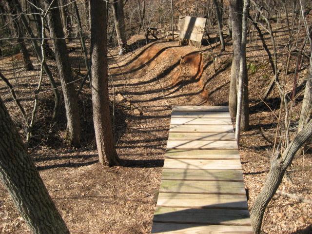 A wooden ramp leading into a dirt jump path in a wooded area, with bare trees surrounding the scene and fallen leaves covering the ground. The jump ramp is built on a slight hillside, creating a path for biking or other outdoor activities. Salem Lake mountain bike trail.