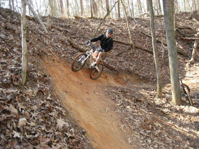 A mountain biker riding down a dirt slope covered in fallen leaves, surrounded by trees in a forested area. The biker is wearing a helmet and a black jacket, demonstrating an action shot as they maneuver the terrain. Salem Lake mountain bike trail.