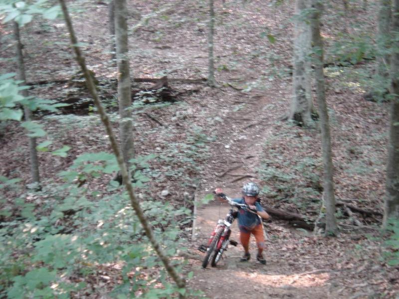 A young cyclist walking uphill on a dirt trail in a wooded area, carrying a bicycle. The scene features trees and leaves in a natural setting, indicating a serene outdoor environment. San-lee Park mountain bike trail.