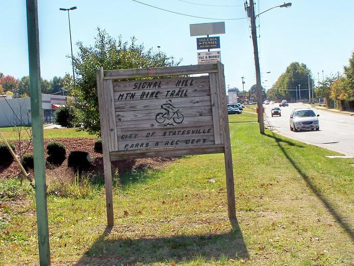 Sign for Signal Hill Mountain Bike Trail in Statesville, NC, displaying a bike symbol and information about the city’s parks and recreation department, with a roadside view and some greenery in the background. Signal Hill mountain bike trail.