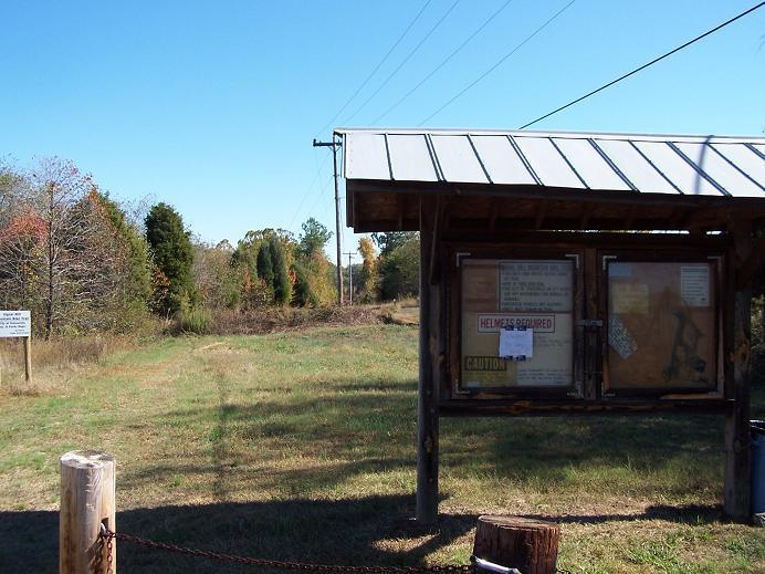 Signage and information board at the entrance of a recreational area, surrounded by tall grass and trees. The clear blue sky is visible above, and power lines run overhead. The board includes safety notices and alerts, with a "Helmets Required" sign prominently displayed. Signal Hill mountain bike trail.