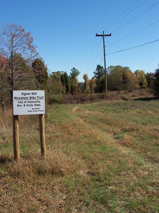 Sign for the Signal Hill Mountain Bike Trail located in Statesville, North Carolina, with a grassy path leading into a wooded area under a clear blue sky. Utility poles are visible in the background. Signal Hill mountain bike trail.