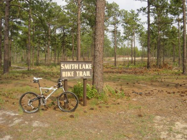 A mountain bike parked next to a wooden sign that reads "Smith Lake Bike Trail," surrounded by tall pine trees and a natural landscape. Smith Lake mountain bike trail.