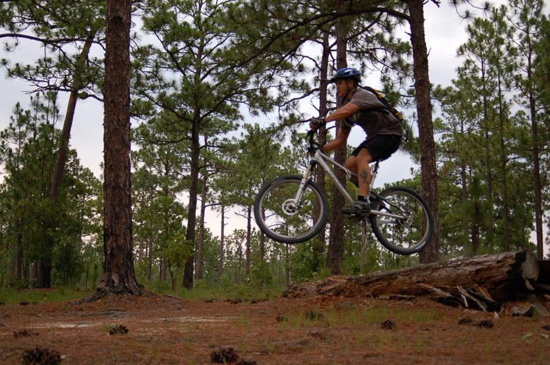A mountain biker jumping over a fallen log in a forested area, surrounded by tall pine trees and a natural, outdoor setting. The cyclist is wearing a helmet and casual biking attire, showcasing an active and adventurous moment. Smith Lake mountain bike trail.