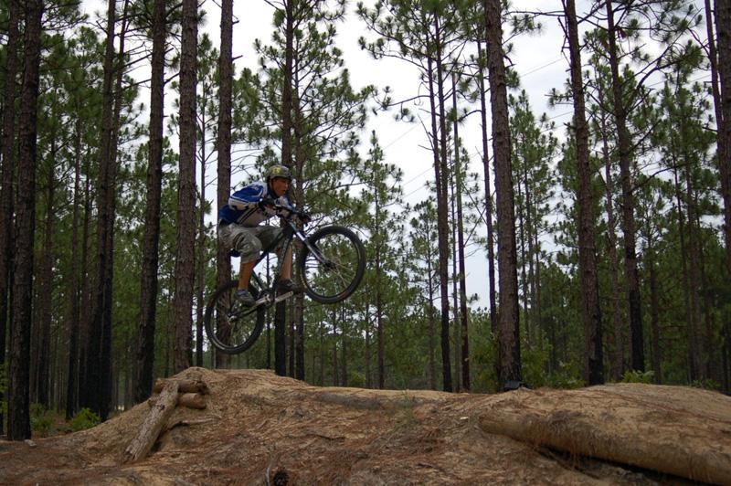 A mountain biker performing a jump over a wooden obstacle on a forest trail surrounded by tall pine trees. The rider is wearing a helmet and a backpack, showcasing an active outdoor scene. Smith Lake mountain bike trail.