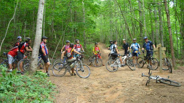A group of cyclists wearing helmets and colorful jerseys stands together along a wooded trail, with their mountain bikes parked nearby. The scene is set in a lush green forest, showcasing trees and a dirt path. Some cyclists are leaning on their bikes while others are chatting, creating a lively atmosphere of camaraderie. Tsali Left Loop mountain bike trail.