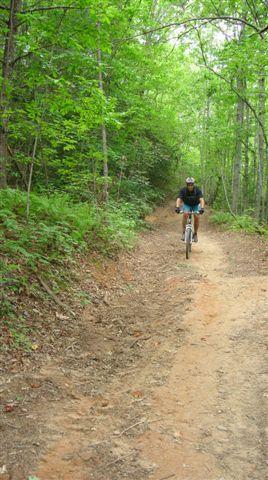 A cyclist riding a mountain bike on a dirt trail through a lush green forest. The path is surrounded by trees and ferns, creating a serene and outdoor atmosphere. Tsali Left Loop mountain bike trail.
