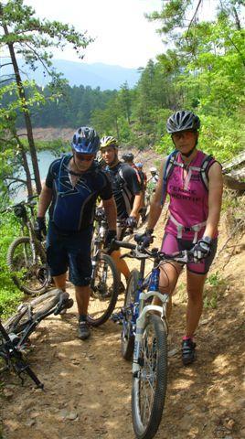 A group of three mountain bikers navigating a dirt trail in a lush green forest. Two are walking beside their bikes, while one is adjusting their bike. The background features trees, mountains, and a glimpse of a body of water. The scene captures the spirit of outdoor adventure and teamwork. Tsali Left Loop mountain bike trail.