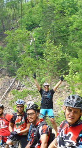 A group of mountain bikers posing for a photo in a wooded area. One cyclist is standing in the back with arms raised in celebration, while others are sitting or standing in the foreground, all wearing bicycle helmets and gear. Green trees and rocky terrain can be seen in the background, indicating an outdoor biking environment. Tsali Left Loop mountain bike trail.