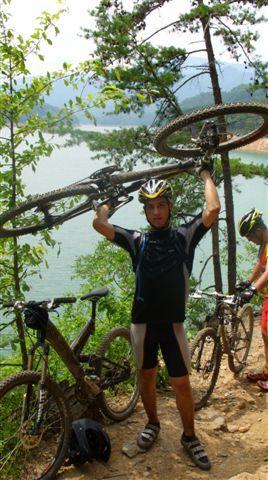 A cyclist in a black and blue jersey raises a mountain bike above his head, standing on a trail surrounded by greenery. In the background, a lake and hills are visible, creating a scenic outdoor setting. Another cyclist is seen nearby, also on a mountain bike. Tsali Left Loop mountain bike trail.