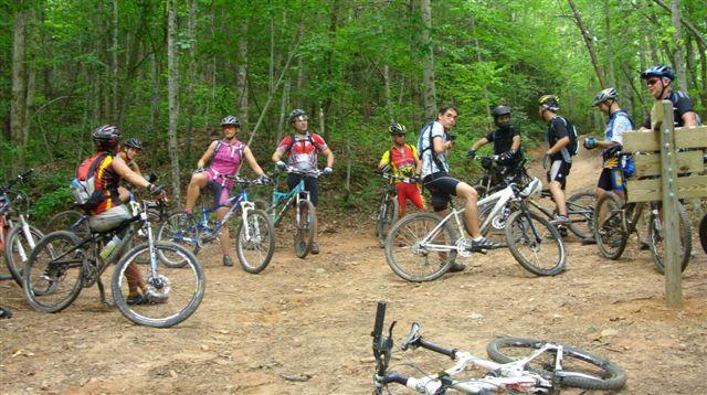 A group of mountain bikers in various riding gear is gathered on a dirt trail surrounded by trees. Some riders are on their bikes, while others are standing or sitting, taking a break. The scene captures a lively outdoor adventure atmosphere, with a mix of bike styles and colorful clothing. In the foreground, a bike is lying on the ground, indicating an active biking environment. Tsali Left Loop mountain bike trail.