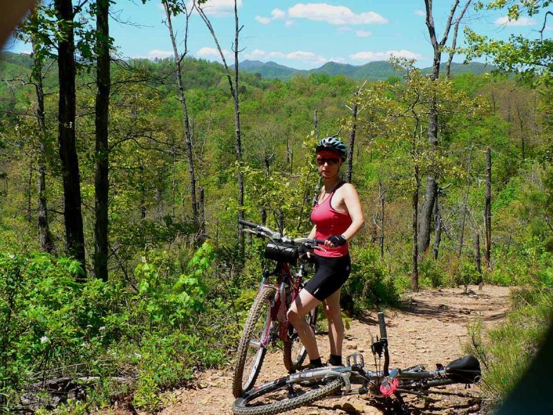 A woman in biking attire stands next to her mountain bike on a dirt trail surrounded by lush green trees and mountains in the background. She is wearing a helmet and sunglasses, with a clear blue sky overhead. The scene depicts an active outdoor lifestyle in a beautiful natural setting. Tsali Left Loop mountain bike trail.