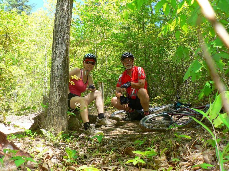 A man and a woman are sitting on a dirt trail surrounded by lush green foliage, both wearing helmets and athletic clothing suitable for biking. The woman is dressed in a pink tank top and shorts, while the man is in a red and black cycling jersey. Two mountain bikes are resting on the ground nearby. The scene is bright and sunny, suggesting an enjoyable outdoor activity. Tsali Left Loop mountain bike trail.