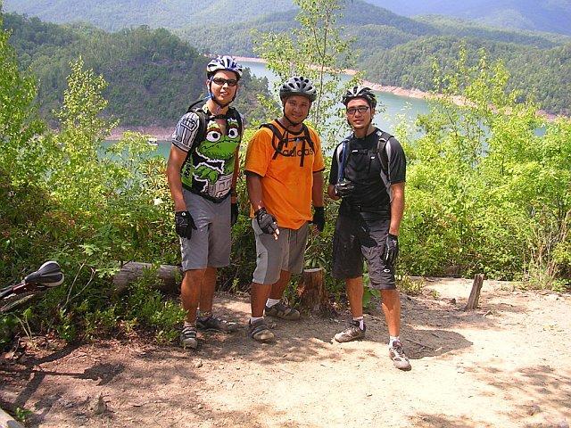Three mountain bikers posing on a trail with a scenic lake and wooded mountains in the background. The first biker wears a colorful shirt featuring a cartoon character, the second is in an orange shirt, and the third is in a black outfit, all wearing helmets and gloves. Tsali Mouse Branch Loop mountain bike trail.