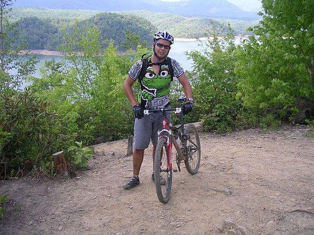 A man standing beside a mountain bike on a dirt path, wearing a helmet and a colorful shirt featuring a character design. The background includes green trees and a scenic view of a lake surrounded by mountains under a partly cloudy sky. Tsali Mouse Branch Loop mountain bike trail.